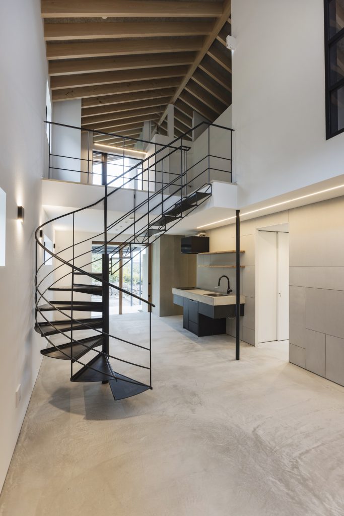 Black steel spiral staircase at Sunset Villa, Awajishima — concrete floor, open-plan kitchen and exposed rafter ceiling beyond