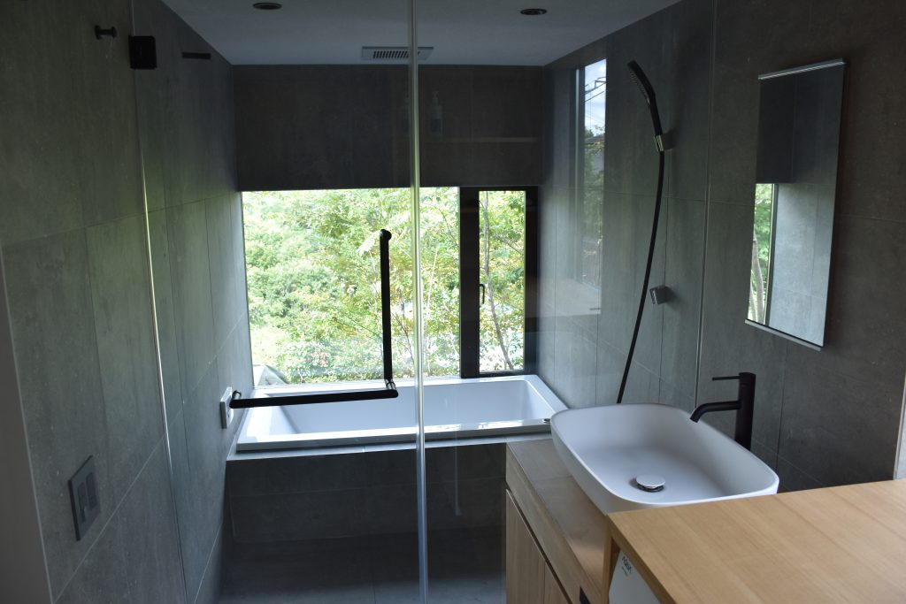 Kato Villa bathroom — vessel basin on timber vanity, freestanding soaking tub, black fixtures and full-height concrete tile finish with garden window