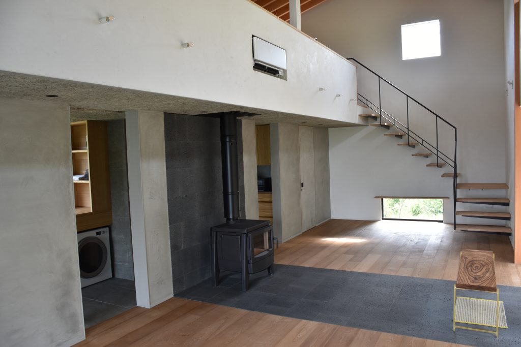 Kato Villa open-plan living area with wood-burning fireplace, concrete walls, timber staircase and double-height void with rooflight