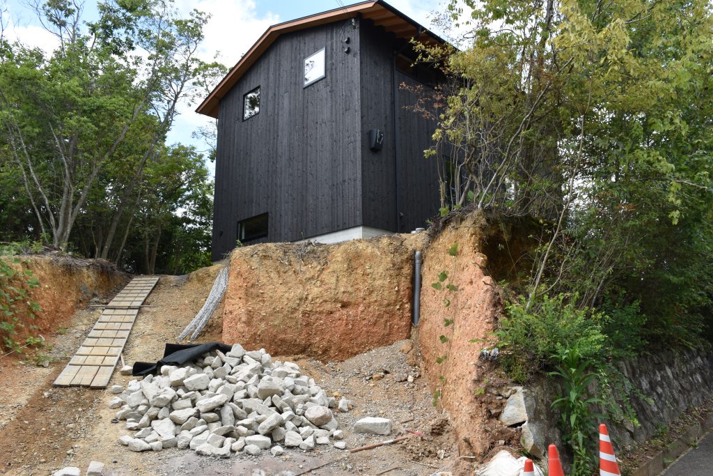 Kato Villa exterior during carport excavation — charred black timber cladding above cut slope with earthworks in progress