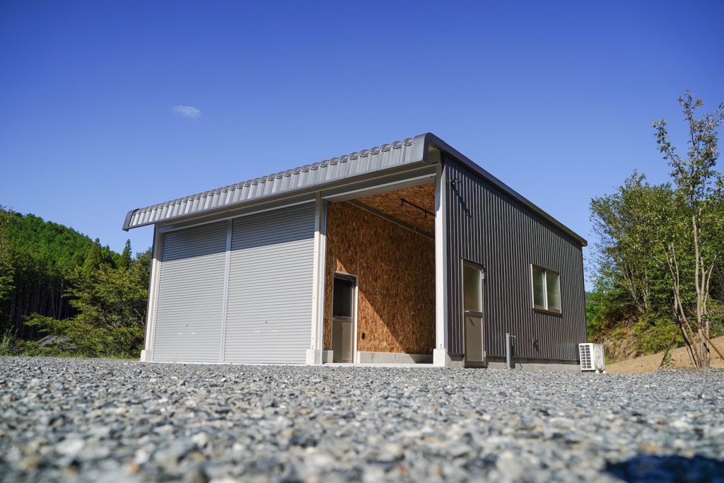 Exterior of a Kakuichi steel garage converted to a clubhouse — corrugated cladding, roller door, and OSB interior visible from forecourt